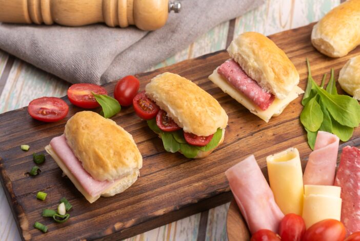 Small sandwiches of ham, salami and cheese with tomato and arugula on a wooden board