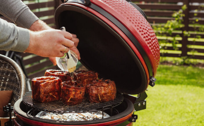 Red Ceramic Barbecue Grill. The man covers the ribs with apple juice. Grilling, Smoking, Baking, BBQ and Roasting process. Quarantine family barbeque in homes terrace.