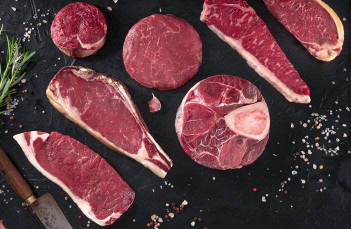 Various cuts of meat, shot from the top on a black background with salt, pepper, rosemary and knives, with copy space