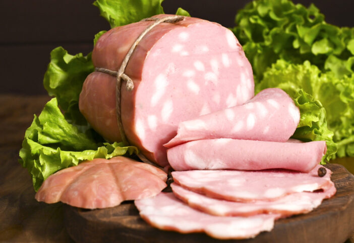 Italian Whole block of mortadella on a wooden plate decorated with green salad. Traditional big sausage and black pepper pistachios Region Emilia Romagna and the city of Bologna. Copy space.