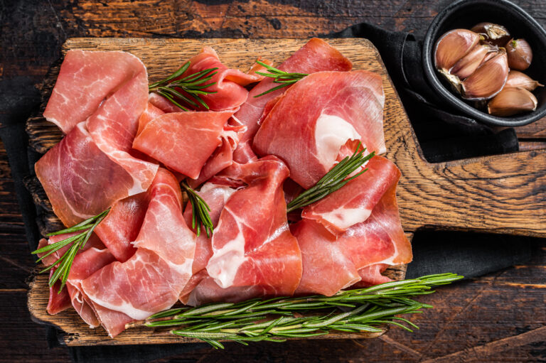 Slices of jamon serrano ham or prosciutto crudo parma on wooden board with rosemary. Wooden background. Top view.