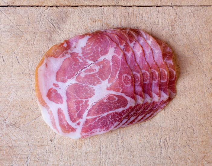 Overhead view of a row of several slices of dry coppa on an old wood cutting board illuminated with natural light.