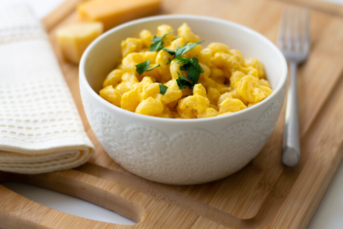 Traditional American macaroni and cheese comfort food (also called mac n cheese) with elbow pasta coated in a cheesy creamy cheddar sauce. Topped with parsley, cheese in the background.