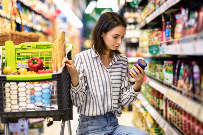 Consumption And Consumerism. Portrait Of Young Woman With Shopping Cart In Market Buying Groceries Food Taking Products From Shelves In Store, Holding Glass Jar Of Sauce, Checking Label Or Expiry Date