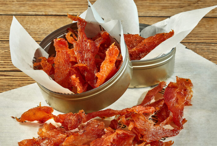 Appetizing spicy dried chicken fillet strips served in tin cans decorated with baking paper on wooden table. Popular meat snack