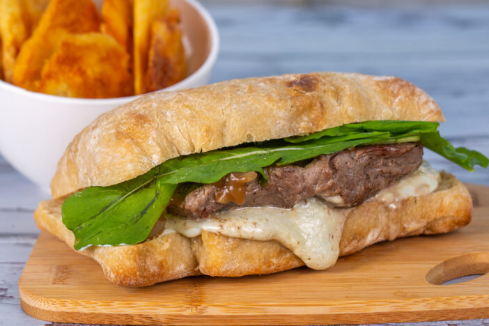 close up meat sandwich with cheese and arugula on ciabatta bread served with potato chips.