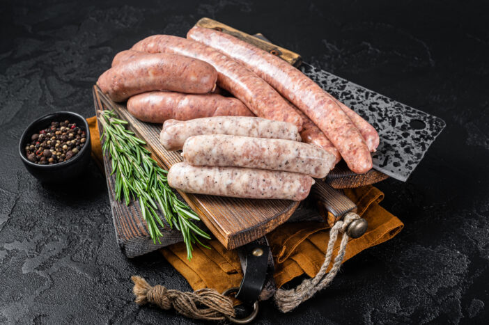 Mix fresh raw sausages. Beef, pork, lamb and chicken mince meat sausages on a butcher cutting board with spices. Black background. Top view.