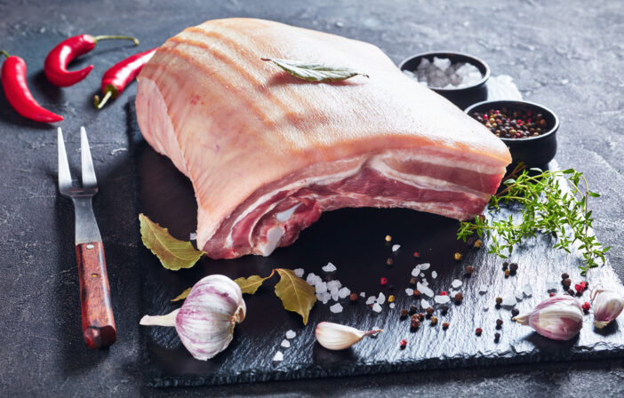 raw pork belly with skin on a black slate tray with spices and herbs, horizontal view from above, close-up