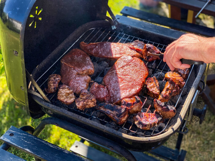 Assorted meat cooking on charcoal kettle grill barbeque. Hand turning meat with tongs. Garden, green grass in background.