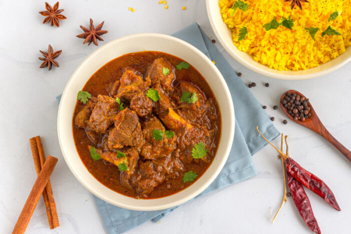 Lamb Curry / Lamb Vindaloo in a Bowl on White Background with Scattered Raw Spices and Colored Rice.