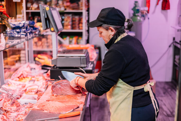 female butcher slicing a beef fillet with a knife on the counter of her butcher shop. In the background, we can see the weighing scale and various fresh products offered in her store. The scene reflects the dedication of the butcher to providing quality cuts and a diverse selection of fresh meat to her customers