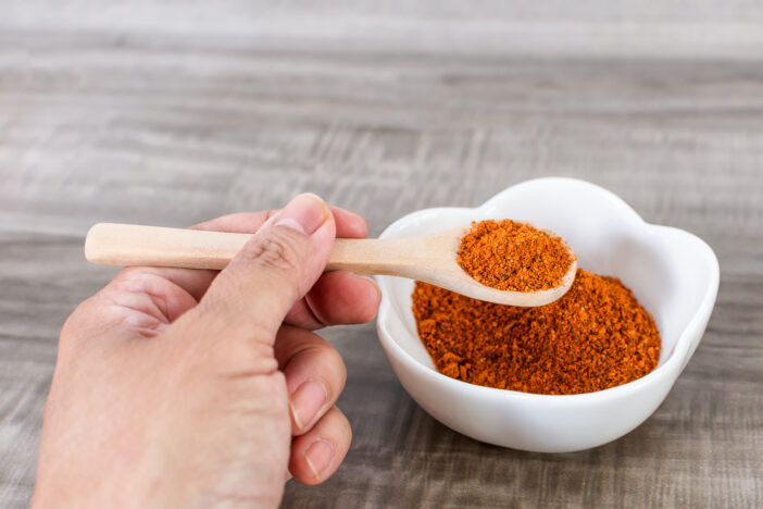 A closeup of a hand holding a wooden spoon full of red pepper