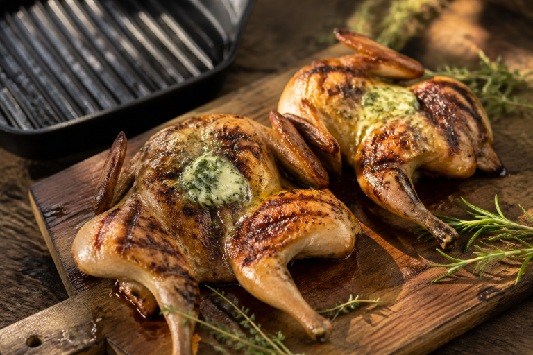 Two grilled butterflied quail on a wooden board topped with melting herb butter and fresh thyme sprigs