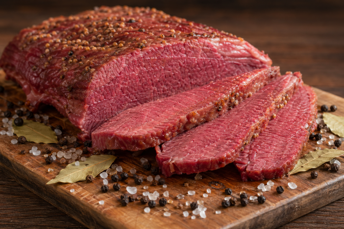 Sliced corned beef brisket on a wooden cutting board surrounded by coarse salt, peppercorns, and bay leaves