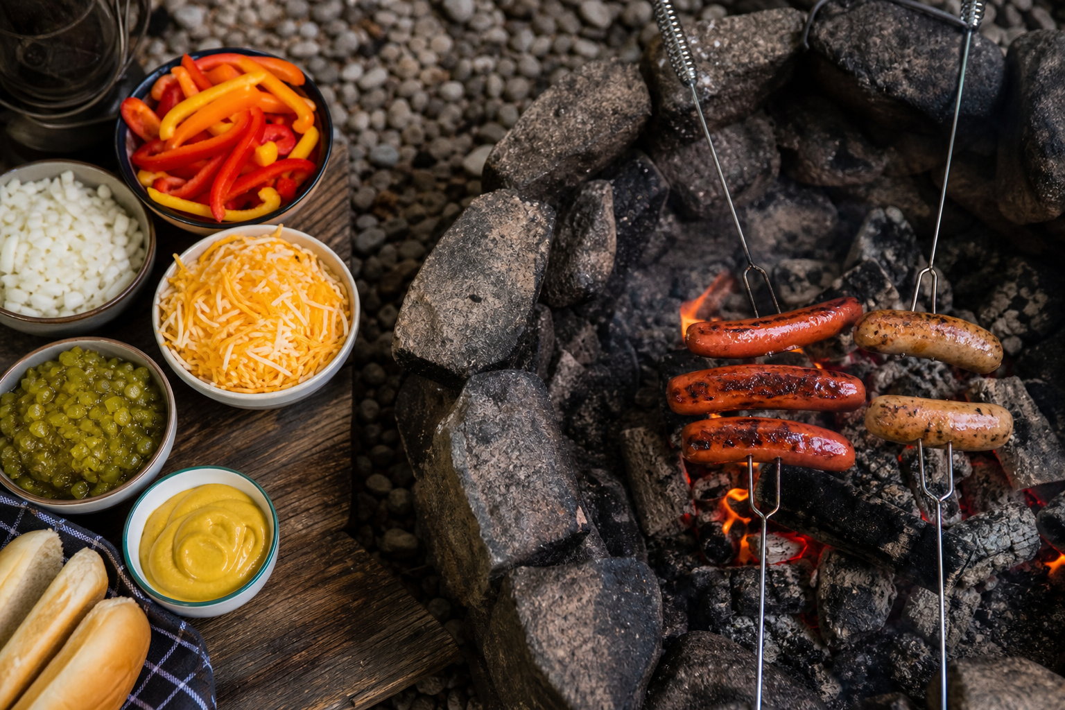 8 Campfire Dinner Ideas Everyone Will Love 17 Assorted sausages and hot dogs on long metal skewers roasting over campfire coals with topping spread nearby